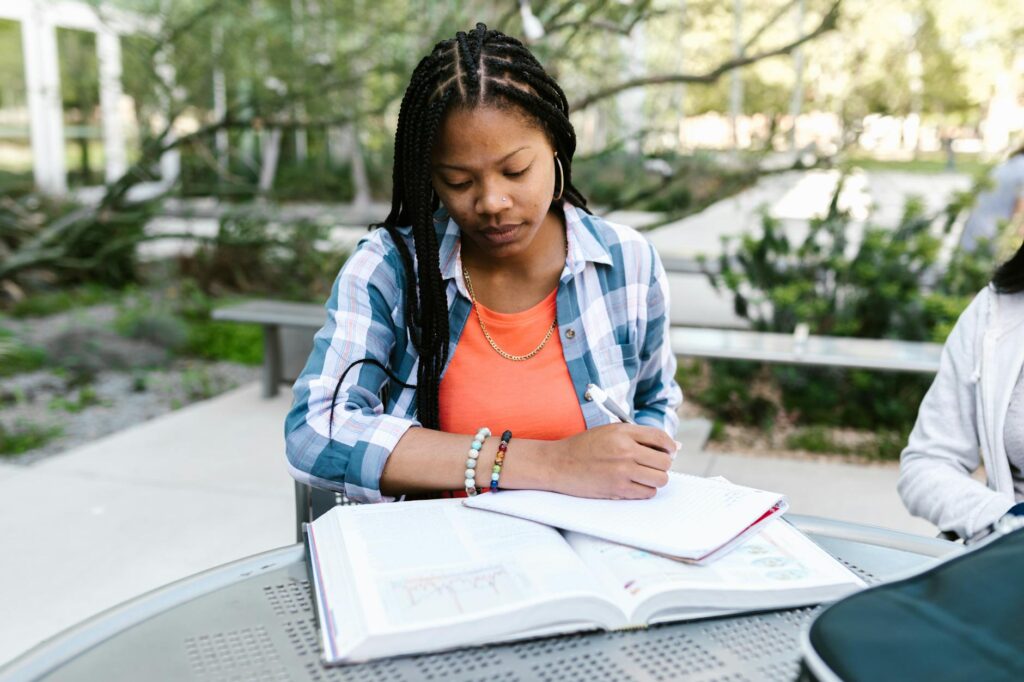 African American woman studying outdoors, writing notes in a notebook.