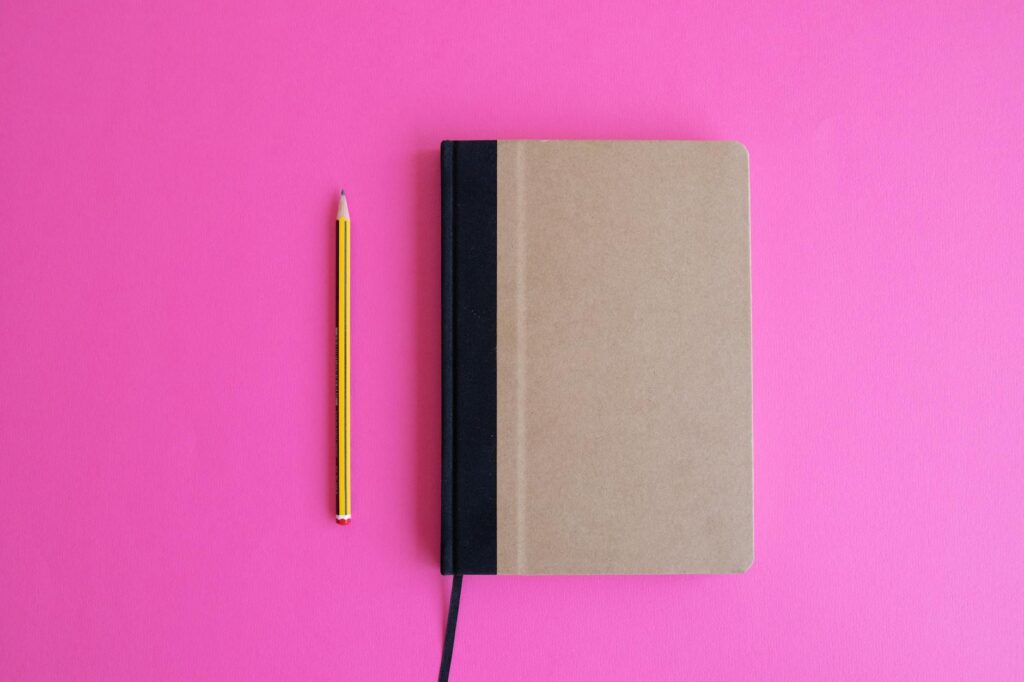 Flat lay of a notebook and pencil placed on a vibrant pink background, perfect for design projects.