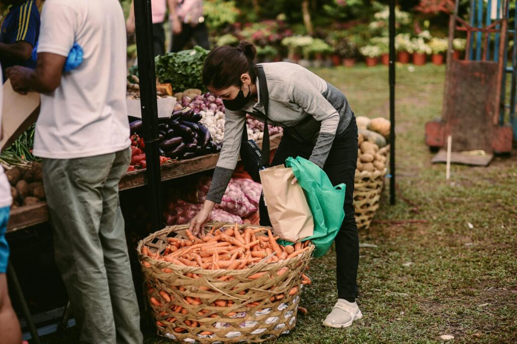 Woman picking fresh carrots at an outdoor vegetable market displaying various produce.