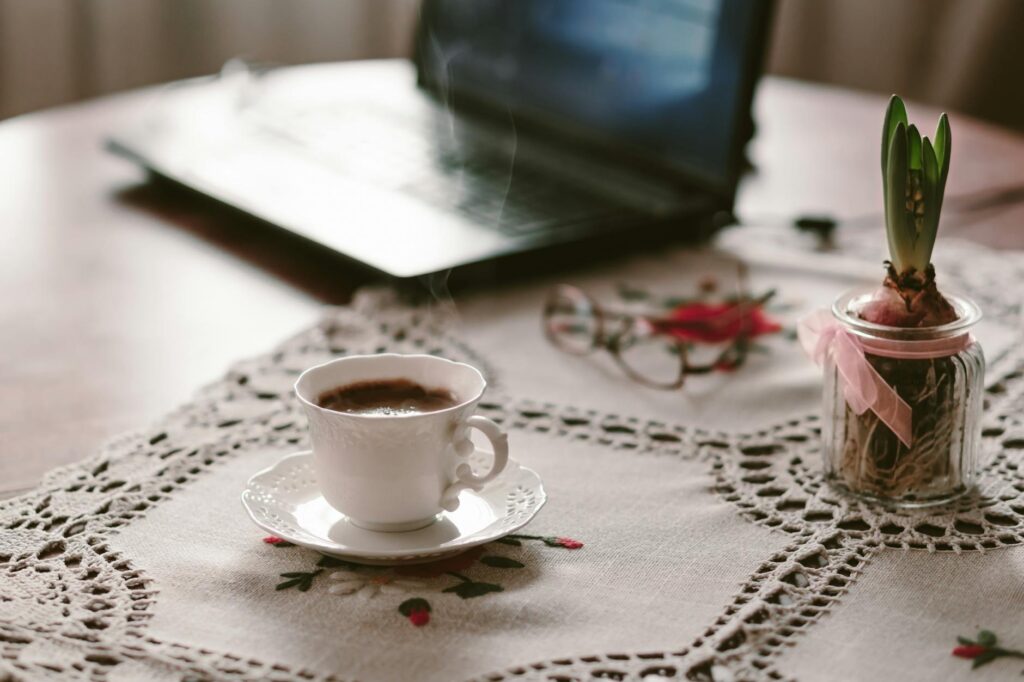 A warm and inviting workspace with a steaming cup of coffee on a lace tablecloth beside a laptop.