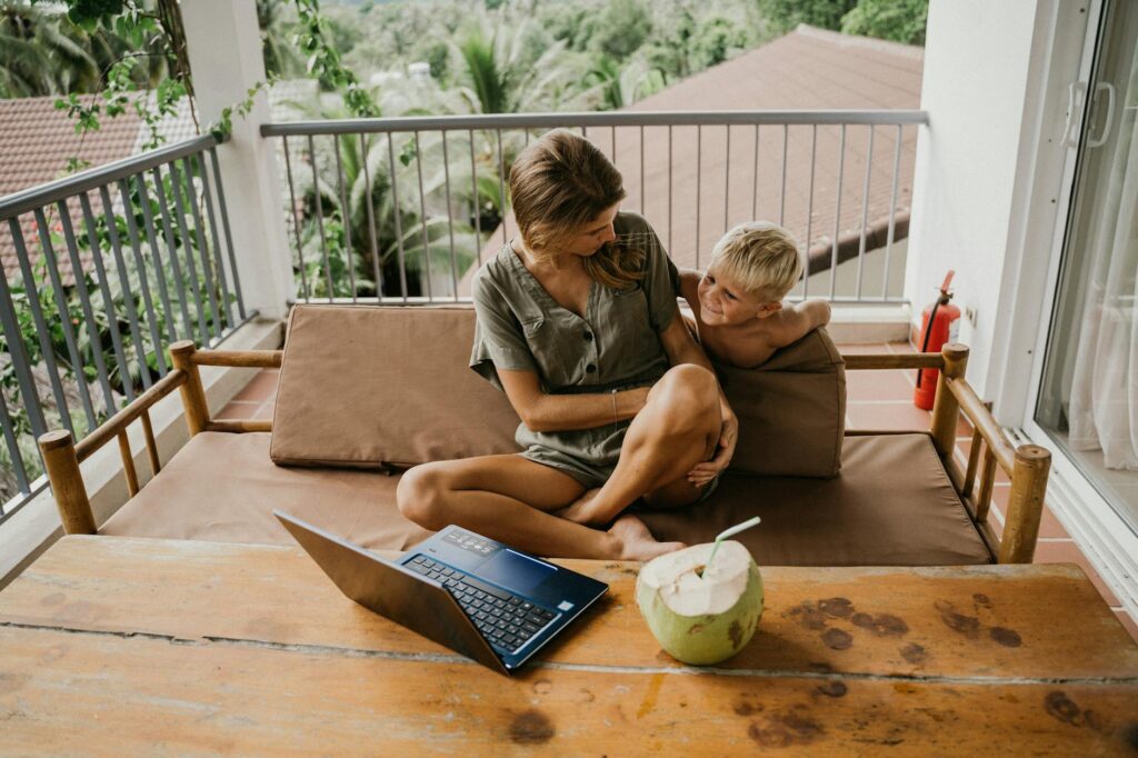 A mother and son relaxing on a balcony with a laptop and coconut nearby.