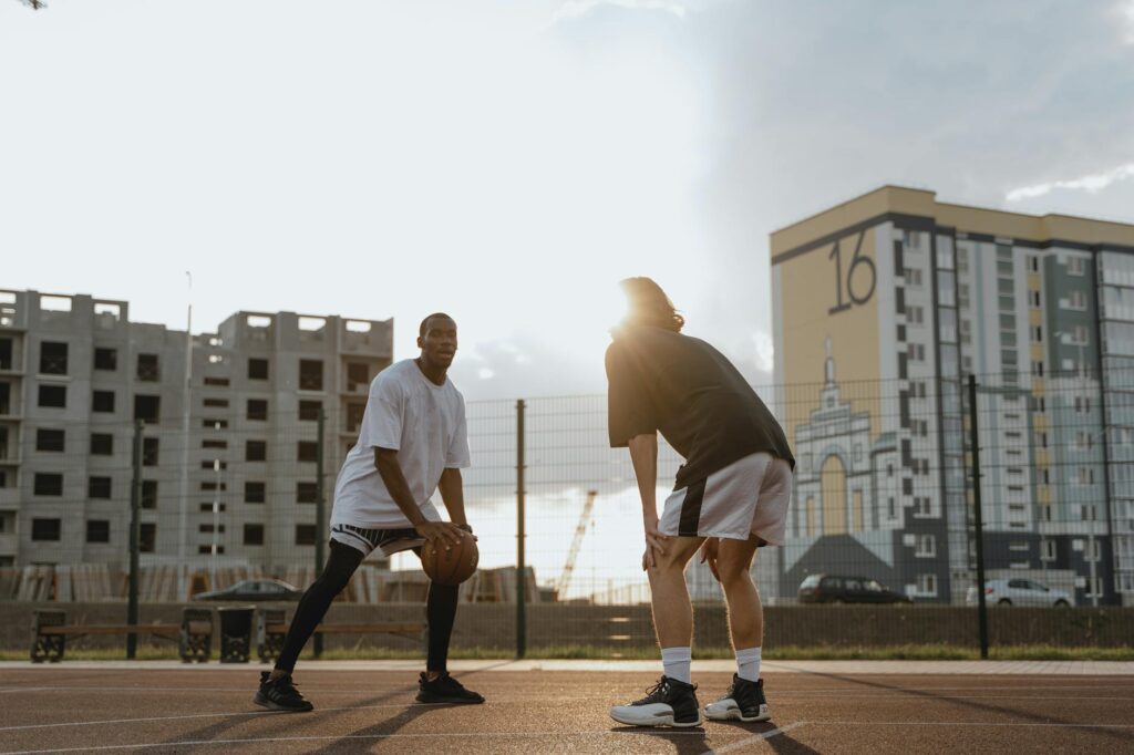 Two young men playing basketball on an outdoor court during sunset near tall buildings.