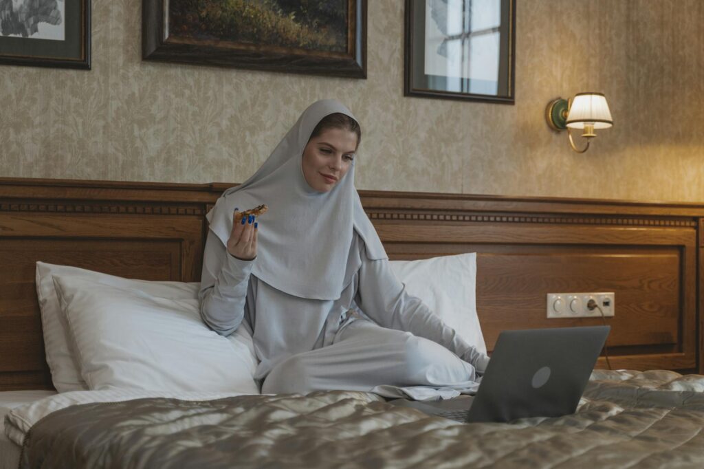 A Muslim woman in a hijab enjoys pizza while watching a laptop in a cozy bedroom setting.