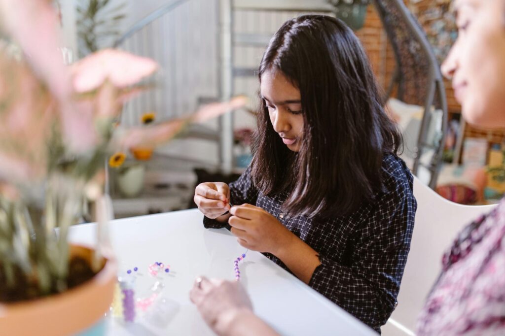 A young girl concentrating on crafting a beaded bracelet at a table indoors.