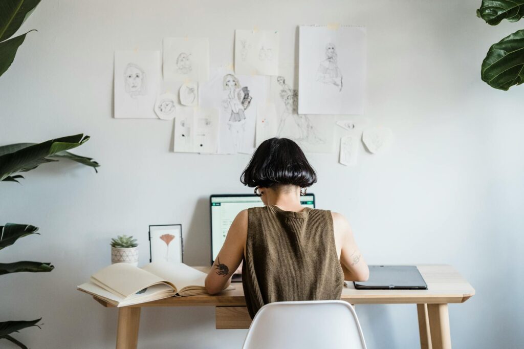 A woman focuses on her laptop in a bright home office adorned with sketches, embodying a creative and freelance lifestyle.