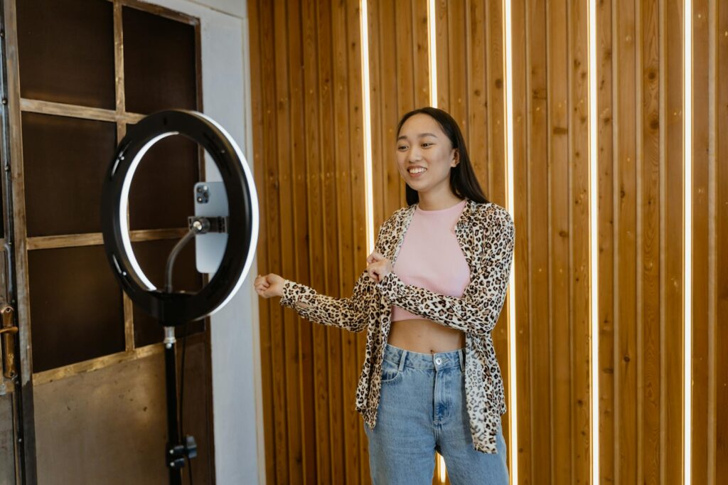 A young woman enjoys dancing indoors with a ring light and smartphone setup, capturing a fun moment.