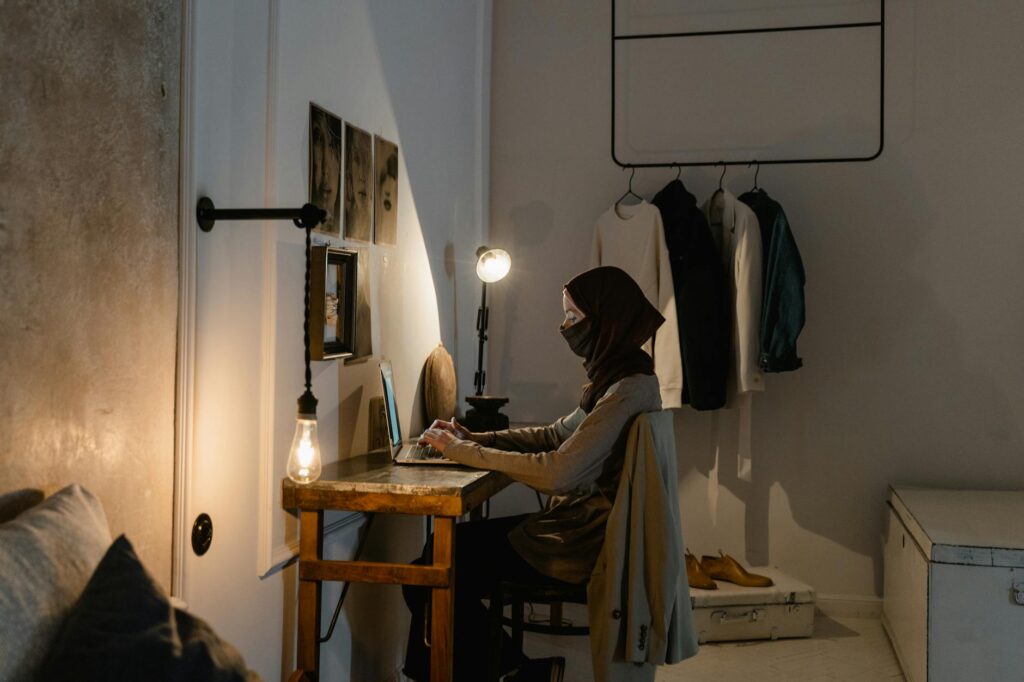 Muslim woman using laptop at home in a cozy, warmly lit workspace with decor.