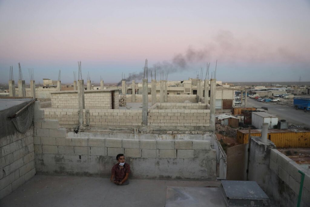 A lone man sits amid unfinished buildings in Syria during twilight, highlighting urban resilience.