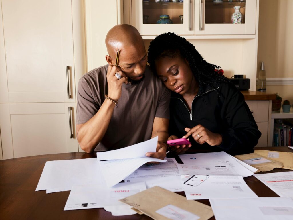 A couple sits at a table managing domestic finances, evaluating documents and using a smartphone.
