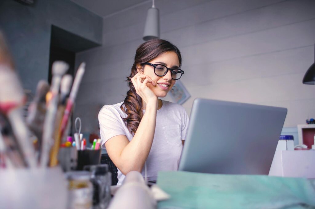 A cheerful woman in glasses works remotely on her laptop in a cozy home office setting.