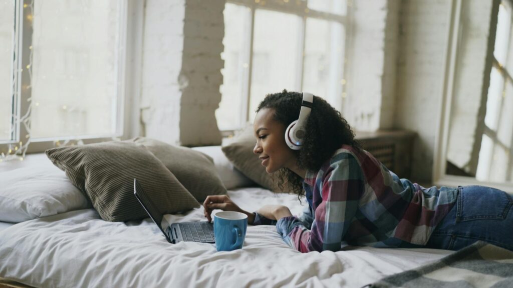 Young woman lounging on bed with headphones, using laptop and enjoying her time.