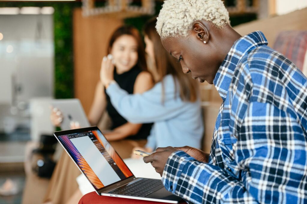 A focused young adult working on a laptop in a collaborative space with coworkers in the background.